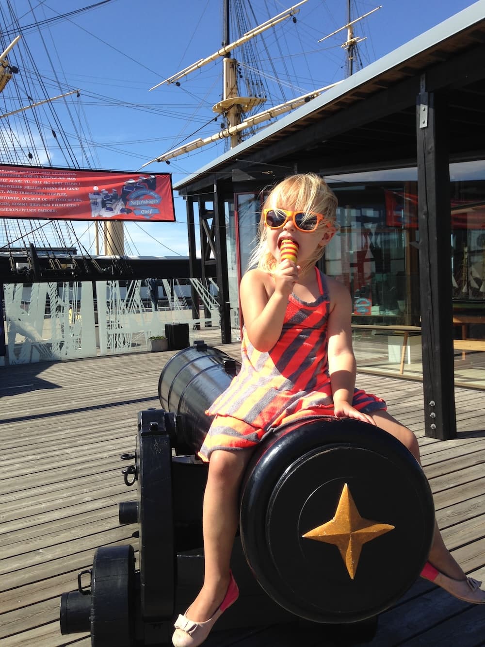 Penelope siddende på en kanon på havnen med sejlskib i baggrunden